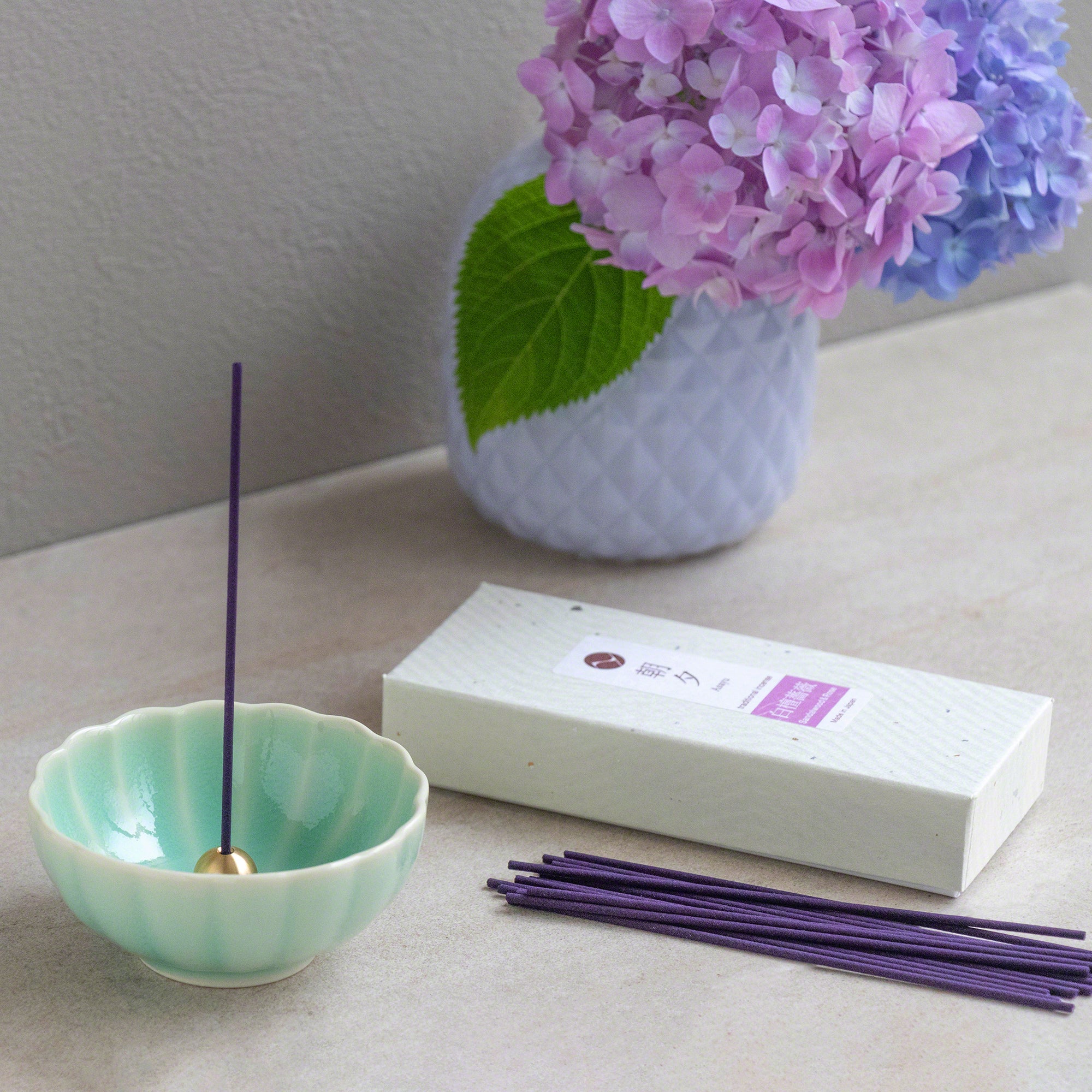 Box of Asayu Japan Sandalwood and Rose Traditional Incense Sticks next to an Asayu Japan Turquoise  Mini Lotus Incense Holder and a vase with violet and pink flowers