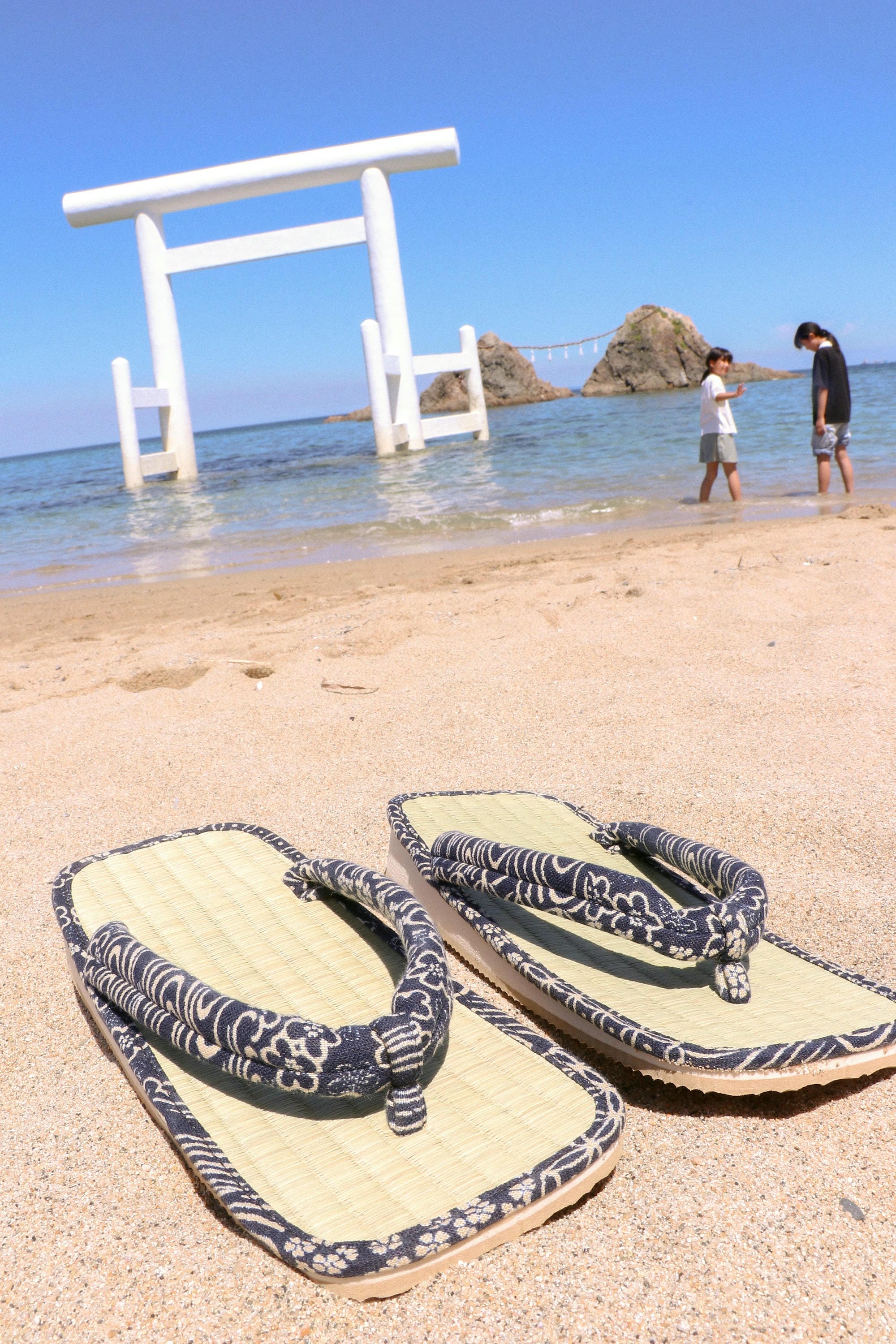 Pair of sandals on a sandy beach with a traditional Japanese torii gate in the background.