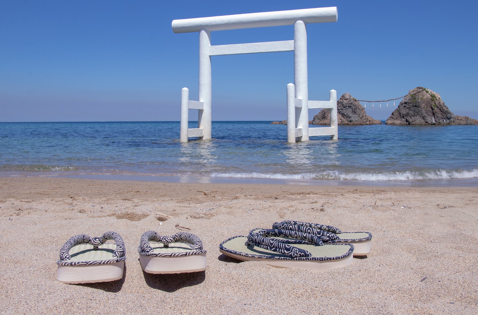 Pair of sandals on a sandy beach with a white torii gate and ocean in the background