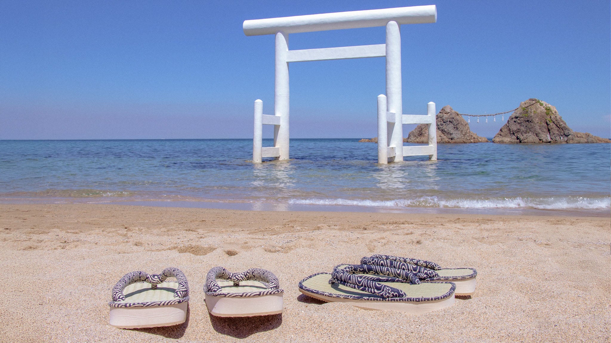 Pair of sandals on a sandy beach in Itoshima with a white torii gate and rock formation in the background.