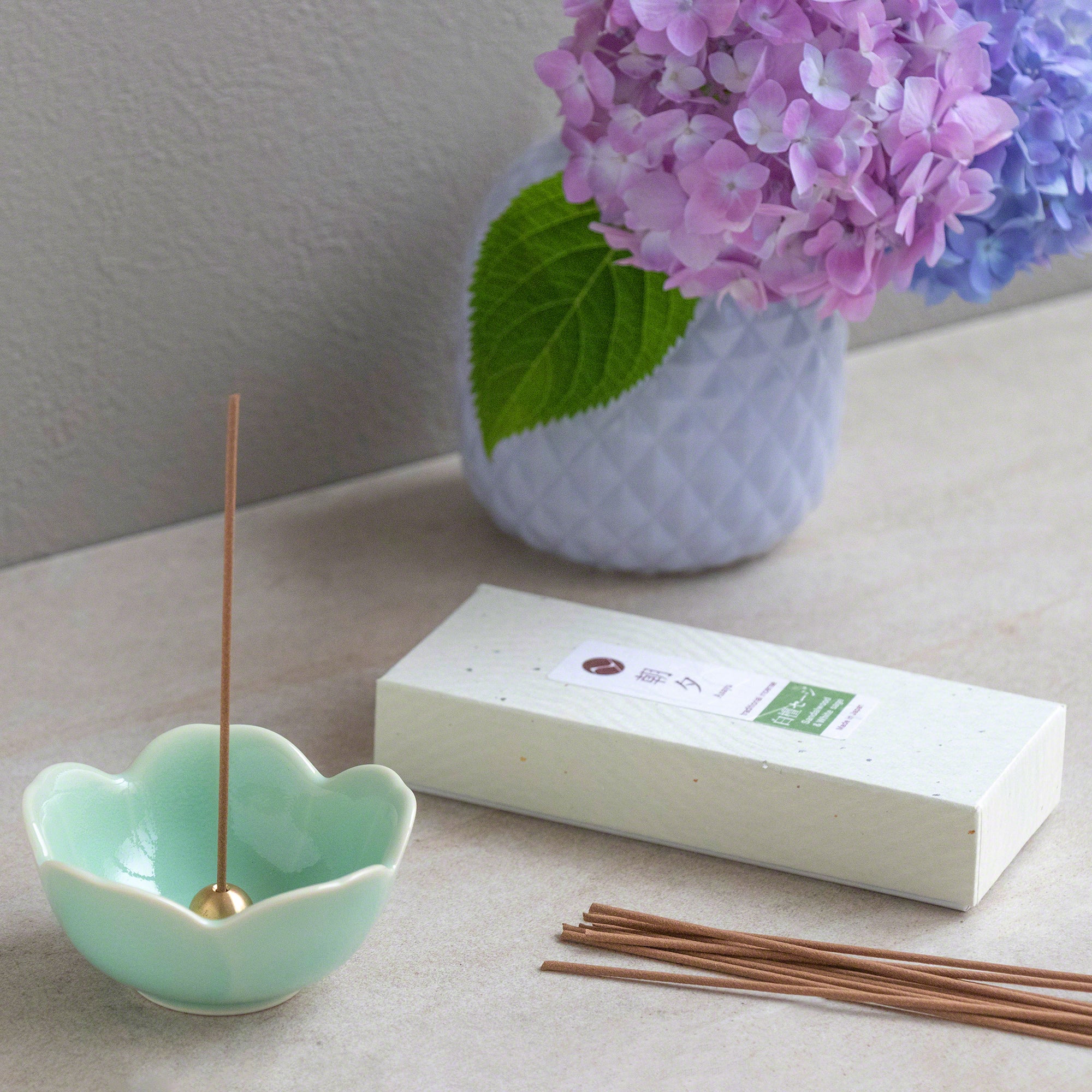 Box of Asayu Japan White Sage and Sandalwood Traditional Incense Sticks next to an Asayu Japan Turquoise Mini Sakura Flower Incense Holder and a vase with violet and pink flowers