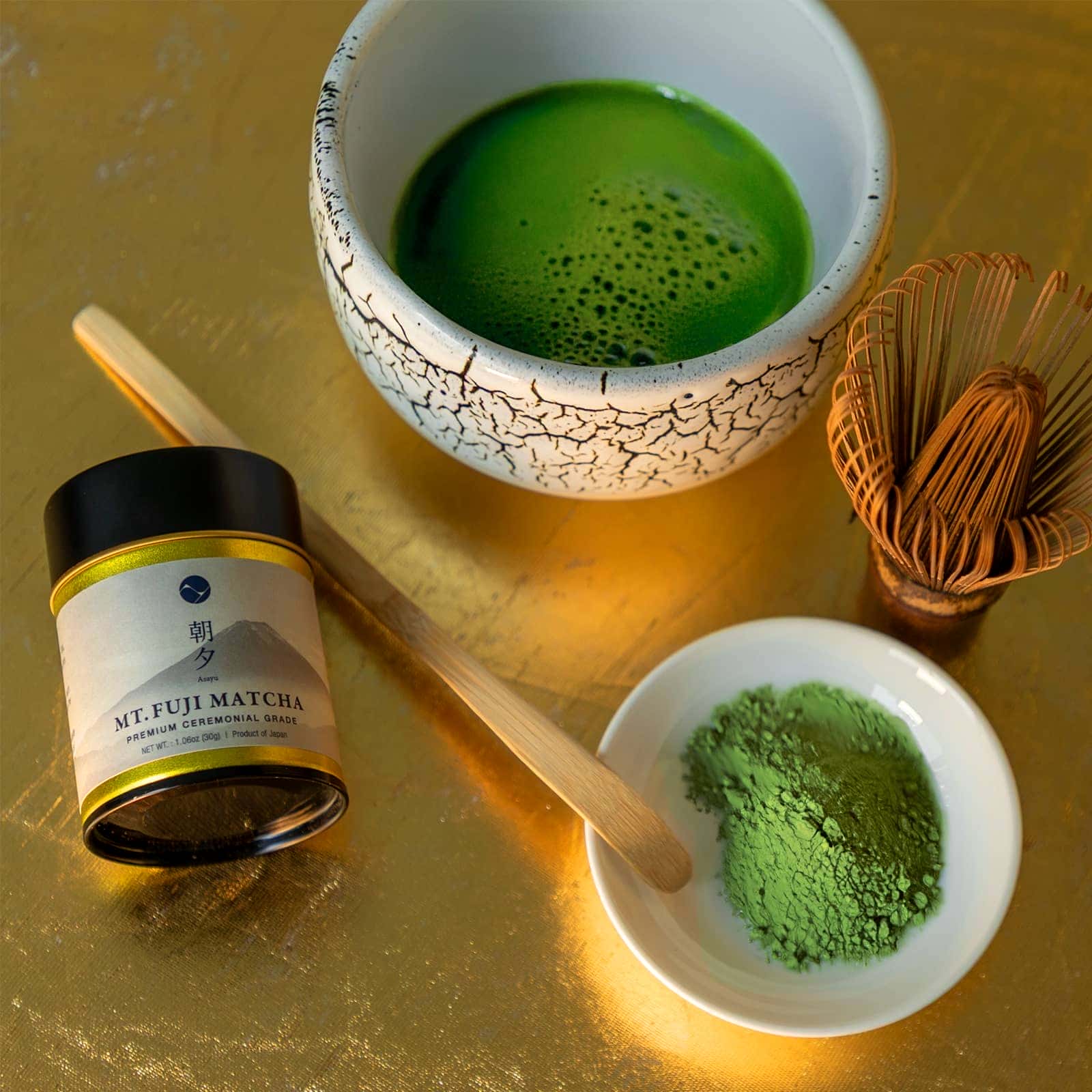 Matcha green tea set with a bowl of matcha, container of matcha powder, and whisk on a gold surface.