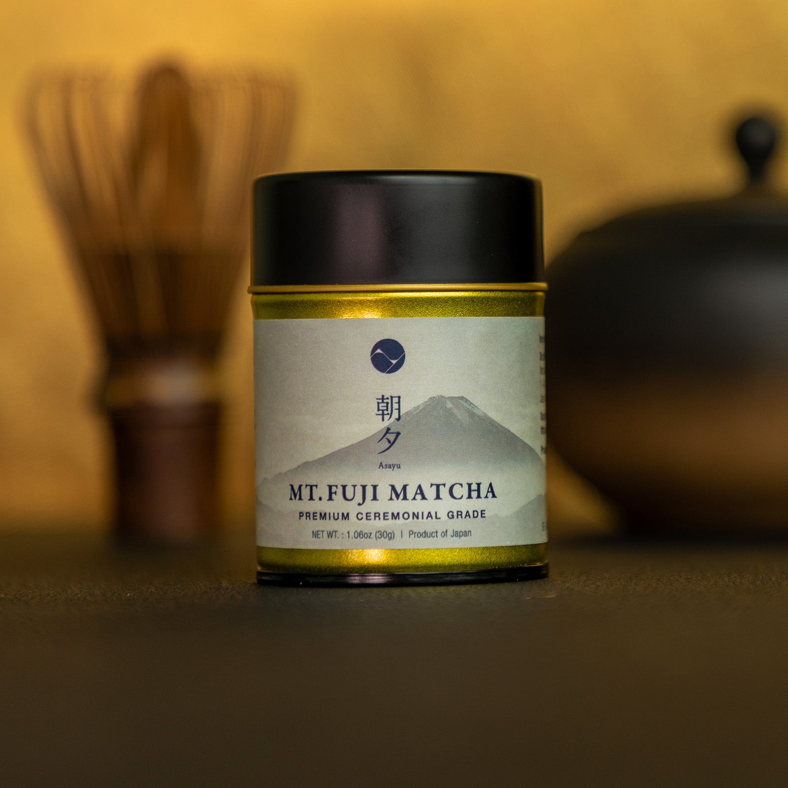Container of Mt. Fuji Matcha with a blurred tea-making setup in the background
