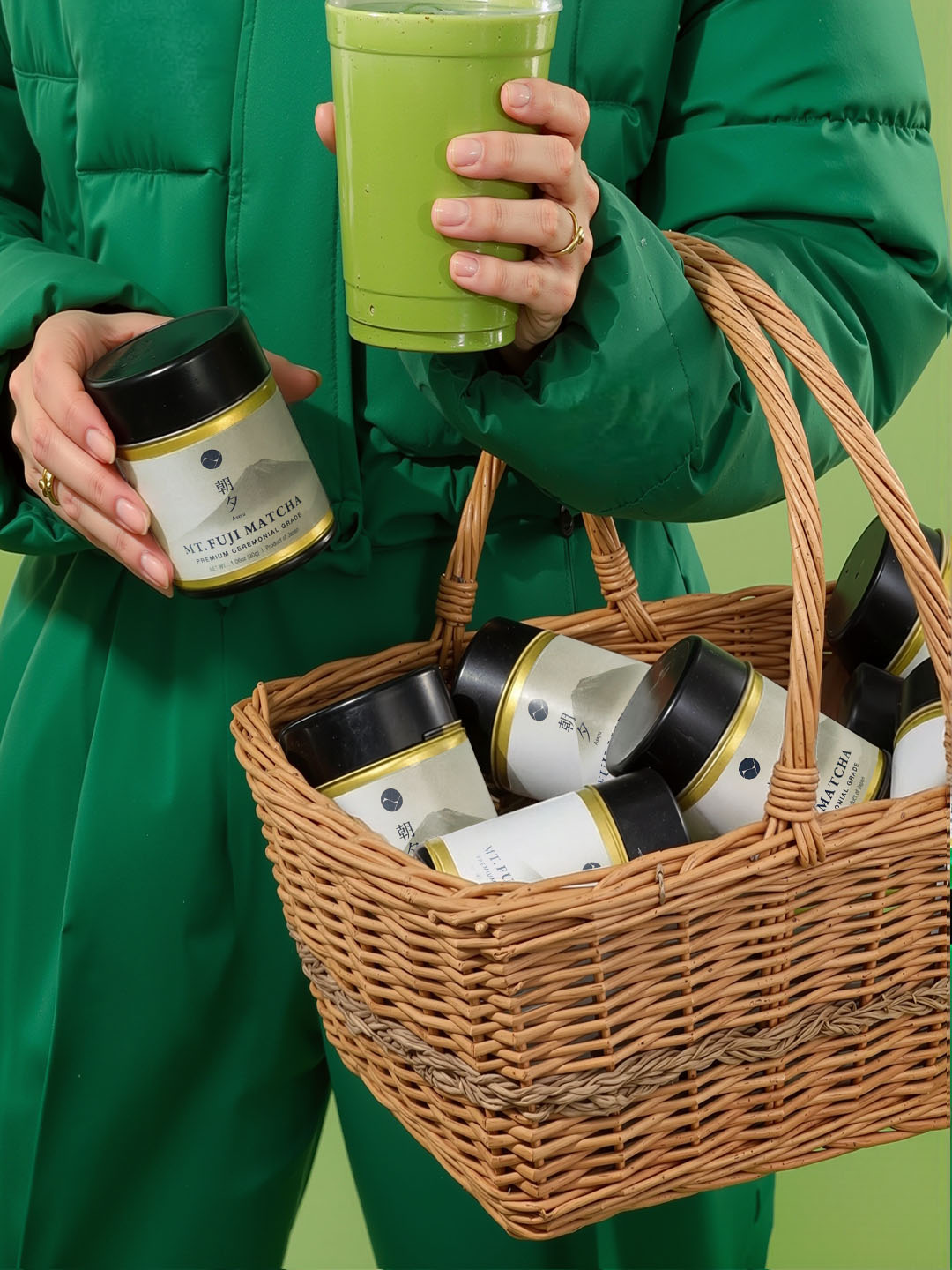 Person holding a green cup and a jar, with a basket containing more jars against a green background