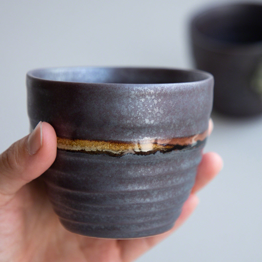 A hand holding one of the Asayu Japan Handpainted Glazed Ceramic Tea Cups Set of 2 in Dark Metallic Brown.