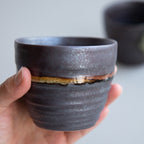 A hand holding one of the Asayu Japan Handpainted Glazed Ceramic Tea Cups Set of 2 in Dark Metallic Brown.