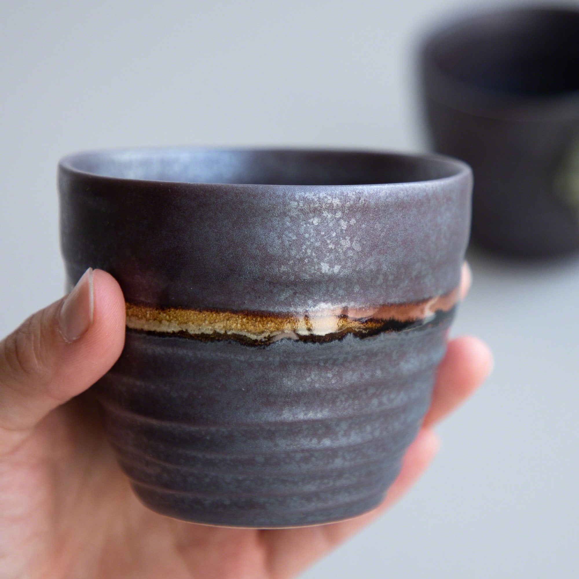 A hand holding one of the Asayu Japan Handpainted Glazed Ceramic Tea Cups Set of 2 in Dark Metallic Brown.