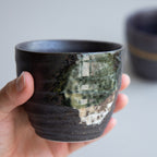 A hand holding one of the Asayu Japan Handpainted Glazed Ceramic Tea Cups Set of 2 in Dark Metallic Brown.