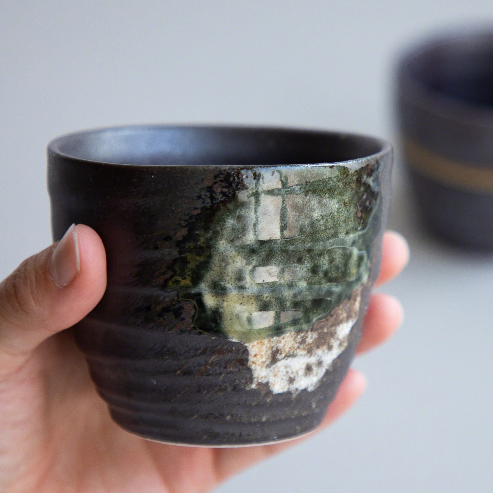 A hand holding one of the Asayu Japan Handpainted Glazed Ceramic Tea Cups Set of 2 in Dark Metallic Brown.