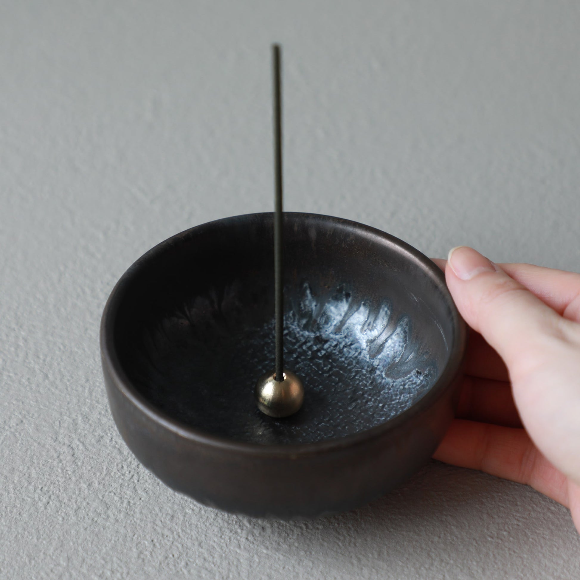 Hand holding Asayu Japan Zen black and gold incense holder with an incense stick on it.