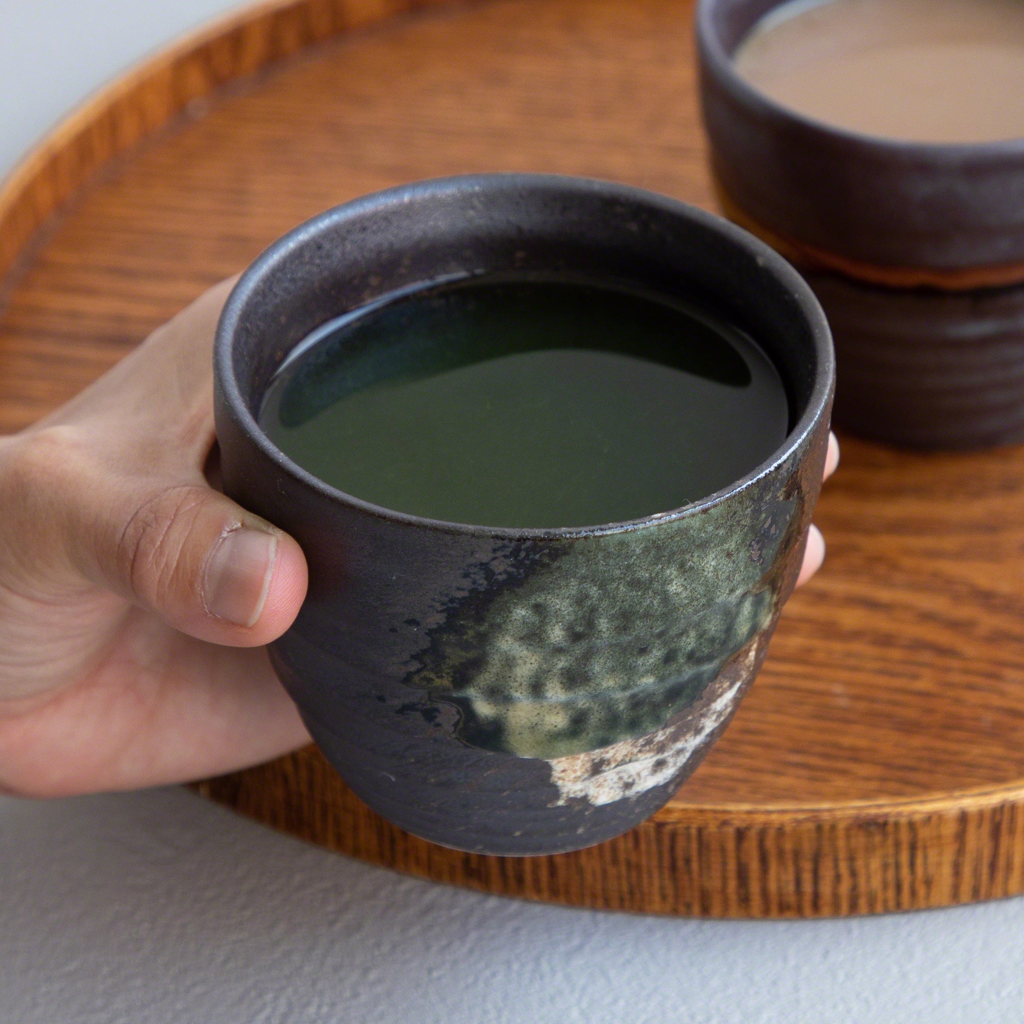 A hand holding one of the Asayu Japan Handpainted Glazed Ceramic Tea Cups Set of 2 in Dark Metallic Brown with green tea.