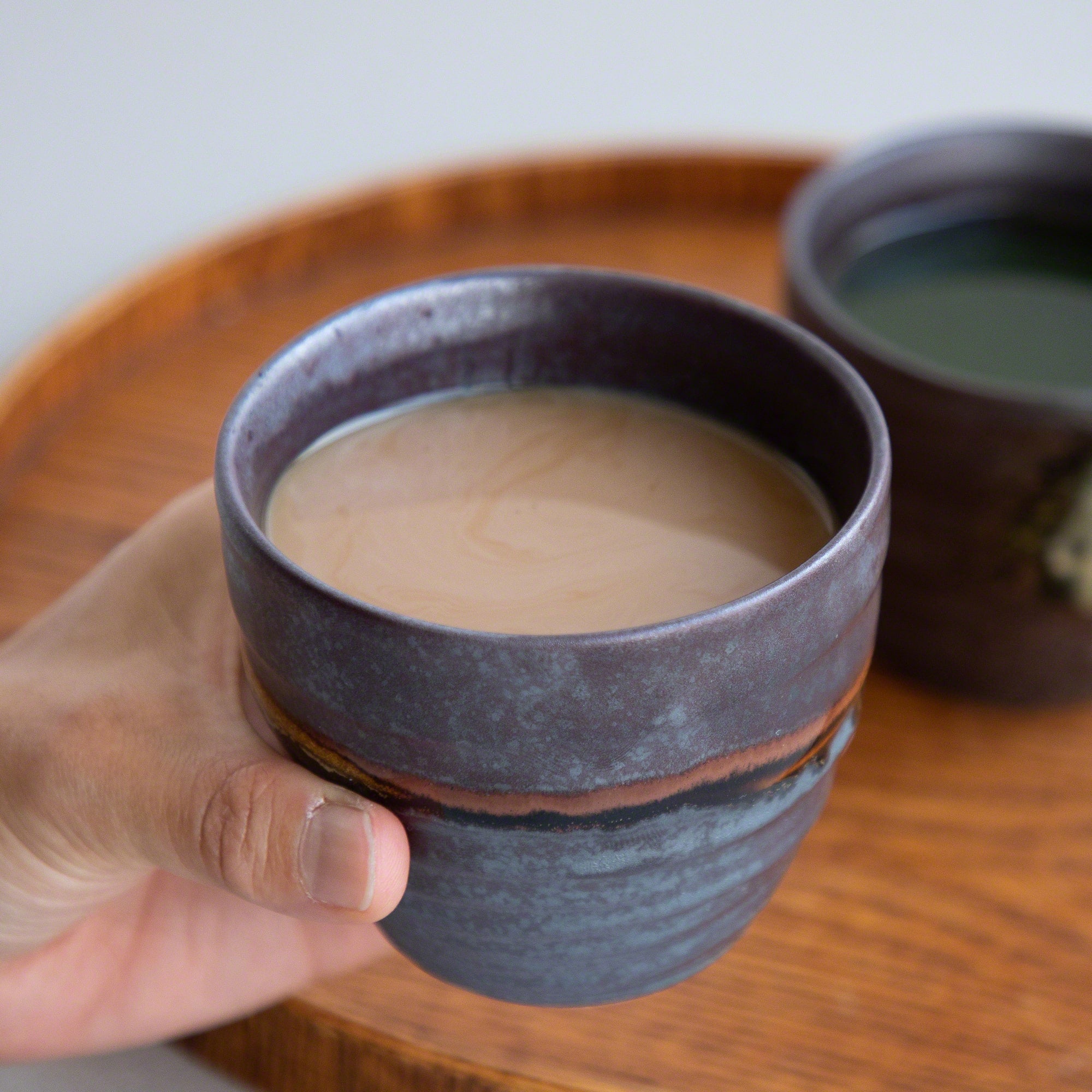 A hand holding one of the Asayu Japan Handpainted Glazed Ceramic Tea Cups Set of 2 in Dark Metallic Brown with Milk Tea.