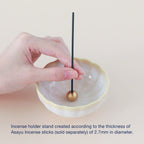 White and yellow ceramic incense holder with brass stand by Asayu Japan with a stick on it.