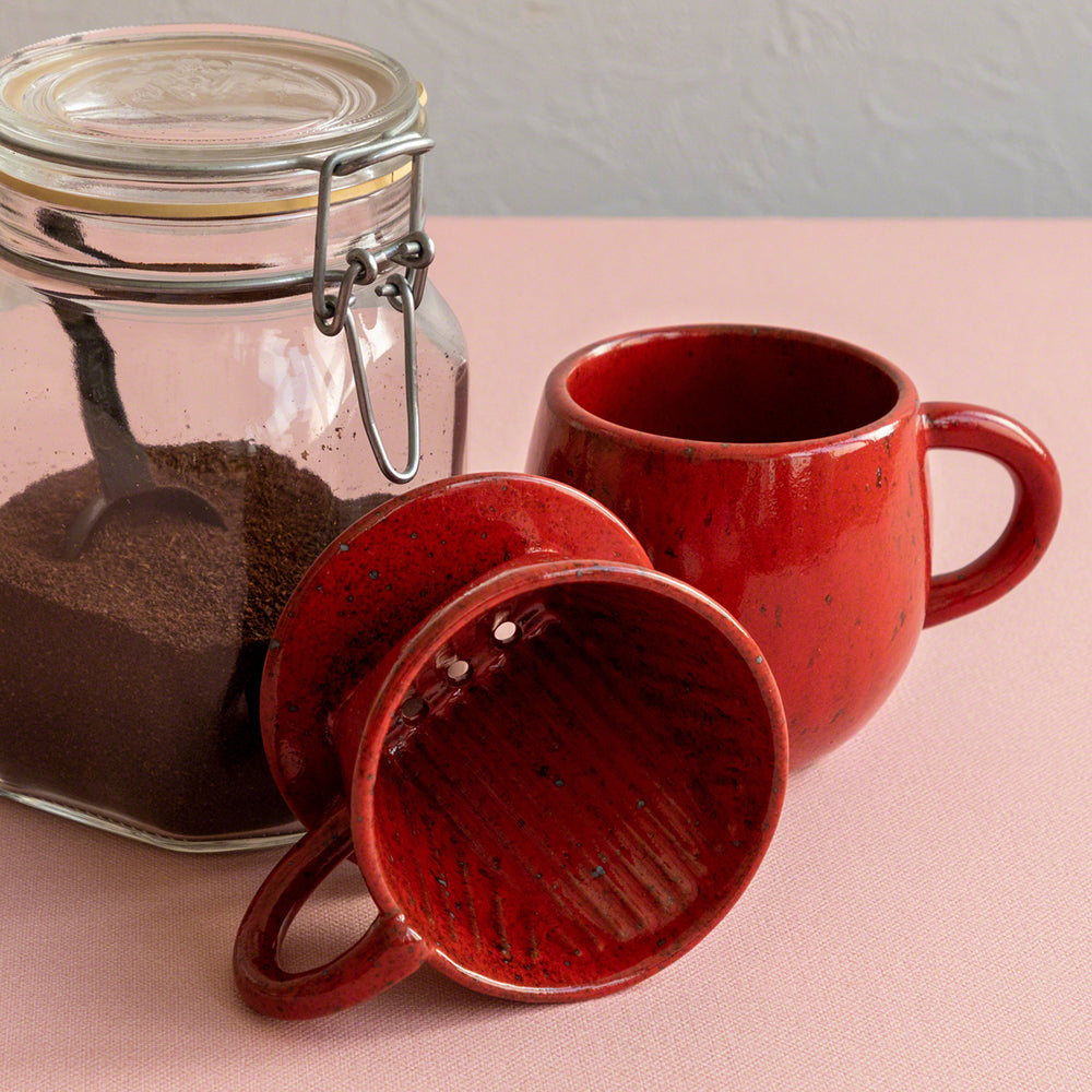 The Asayu Japan Coffee Dripper in chrome red on the side next to the matching accesory mug and a glass container with ground coffee.